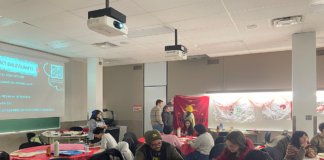 ‘Love as resistance’: Students write letters to political prisoners in the Philippines About a dozen people stand around or sit at several tables decorated with table cloths and filled with papers and writing materials. Some people talk to each other, some are writing on their papers.