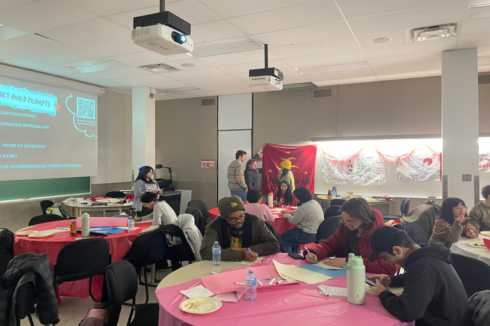 About a dozen people stand around or sit at several tables decorated with table cloths and filled with papers and writing materials. Some people talk to each other, some are writing on their papers.