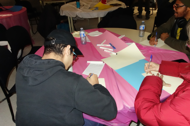 Three people sit at a table with a pink tablecloth that has markers and paper scattered on it.