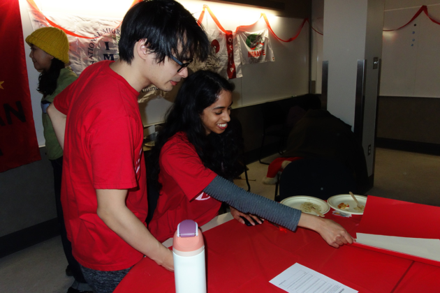 Two people stand in front of a table with writing materials and paper on it. One reaches across the table.