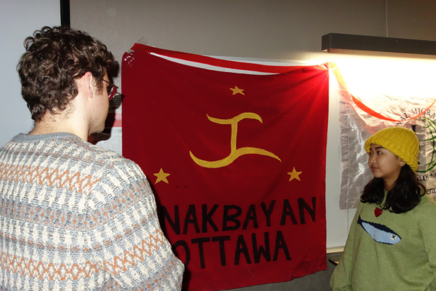 Two people stand and look at a banner that says "Anakbayan Ottawa"