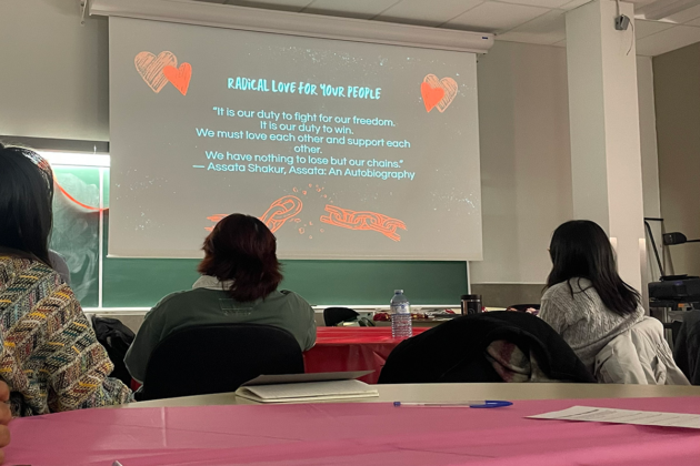 People sit at tables and look up at a projected screen with a powerpoint slide open. The slide says "Radical love for your people: 'It is our duty to fight for our freedom, it is our duty to win, we must love each other and support each other, we have nothing to lose but our chains' - Assata Shakur, Assata: An Autobiography"