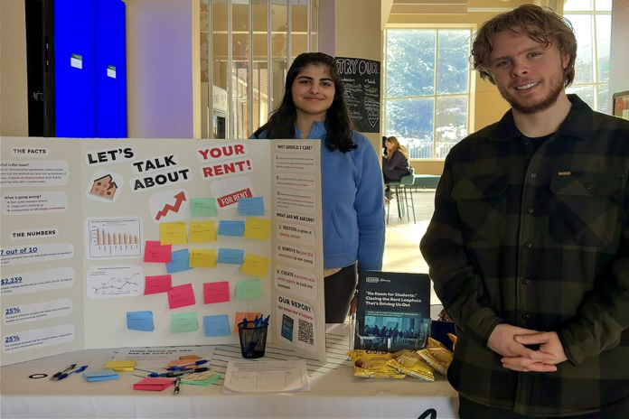 Two people stand at a table with a fold-out cardboard poster with sticky notes and a message that says 