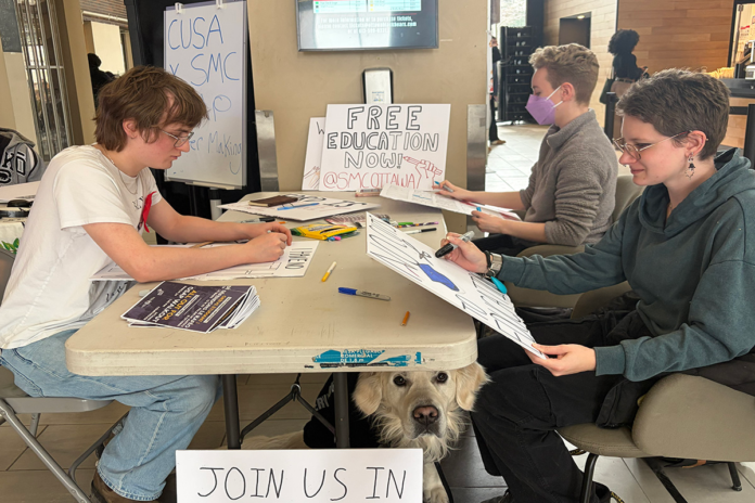 Three people sit at a table that has markers and posters (some homemade, some printed)