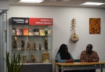 Carleton weaves knowledge with culture through partnership with African universities Two people sit at a tablewith a glass case behind them with statues and books and a sign that says "Carleton University Institute of African Studies"