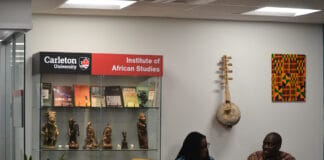 Carleton weaves knowledge with culture through partnership with African universities Two people sit at a tablewith a glass case behind them with statues and books and a sign that says "Carleton University Institute of African Studies"