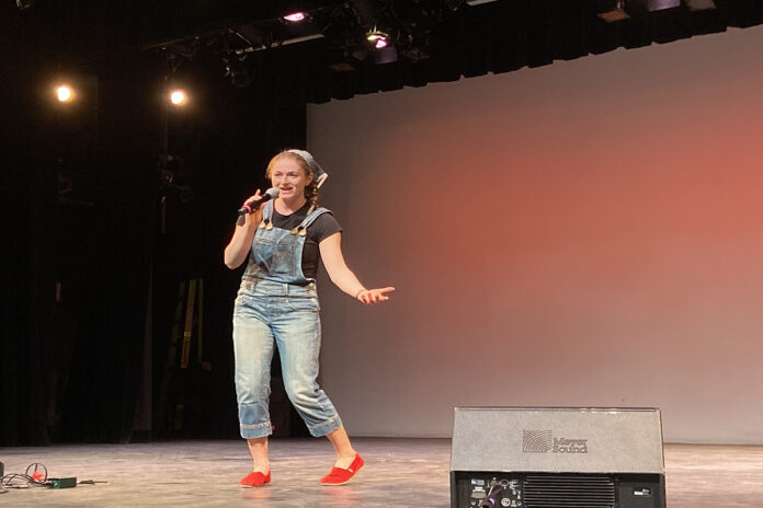 A woman wearing overalls holds a microphone up to her mouth as she stands on a stage.