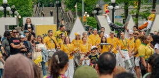 Ottawa’s largest Brazilian festival showcases cultural traditions and community spirit image displaying a crowd a people mostly dressed in yellow smiling and holding drums in front of an Ottawa sign in Ottawa.