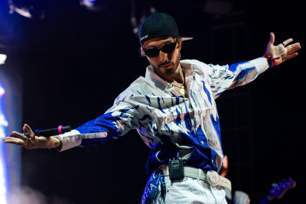 Man outstretches arms on stage, wearing backwards hat and sunglasses