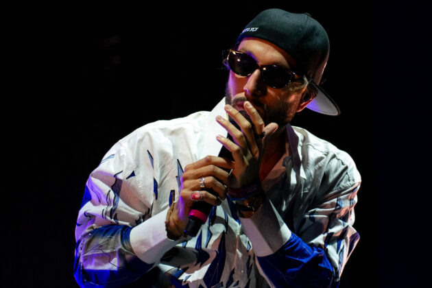 Man holds mic with both hands while singing on stage in sunglasses, backwards hat