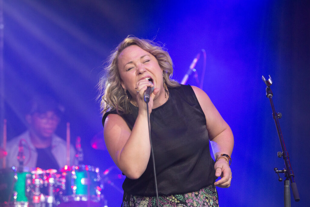 Woman sings into mic with man drumming behind her
