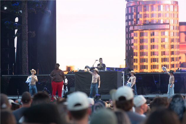 Shirtless boys wave shirts above their head on stage with a rapper in a red tracksuit