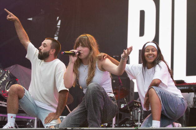 Woman bends down while singing accompanied by two people posing on each side of her