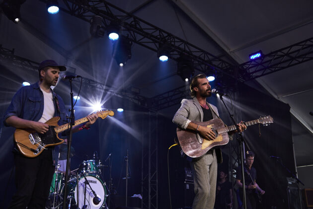 Two men sing at standing mics while each playing guitar