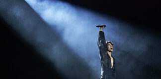Bluesfest Day 5 review: Conan Gray dazzles with dreamy tunes and pop perfection Man wearing black outfit stands in bright spotlight raising microphone to air