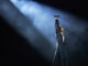 Bluesfest Day 5 review: Conan Gray dazzles with dreamy tunes and pop perfection Man wearing black outfit stands in bright spotlight raising microphone to air