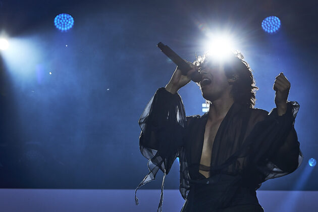 Man is highlighted by bright white stage lights, holds microphone and raises other hands while singing on stage
