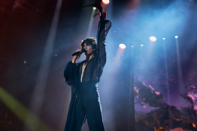 Man stares at crowd while singing into mic with hand raised in air on stage.