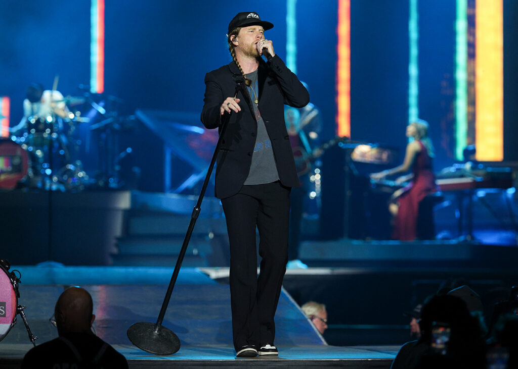 Singer holds microphone stand sideways, holds mic while singing on stage lit by blue.