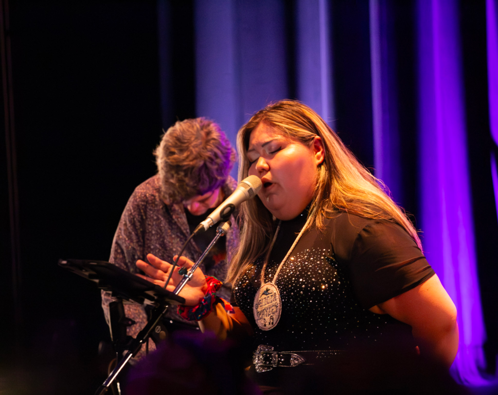 Woman leans over while singing with man playing instrument behind her