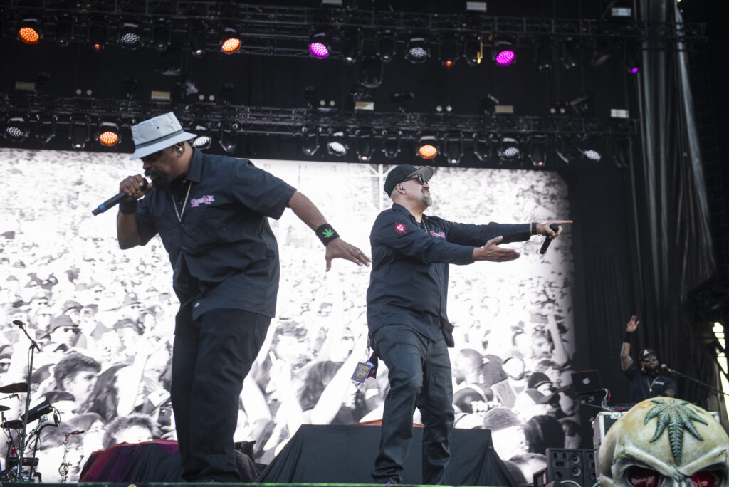 Two men raise mics and dance on stage with bright graphics behind them