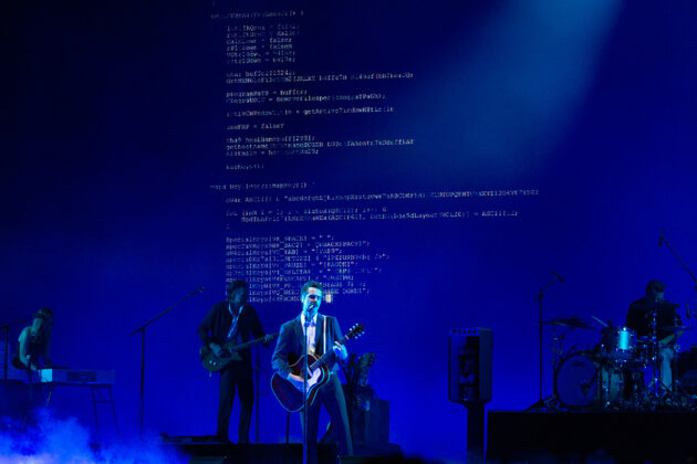 Man sings on stage with surrounding band members in blue hazy light